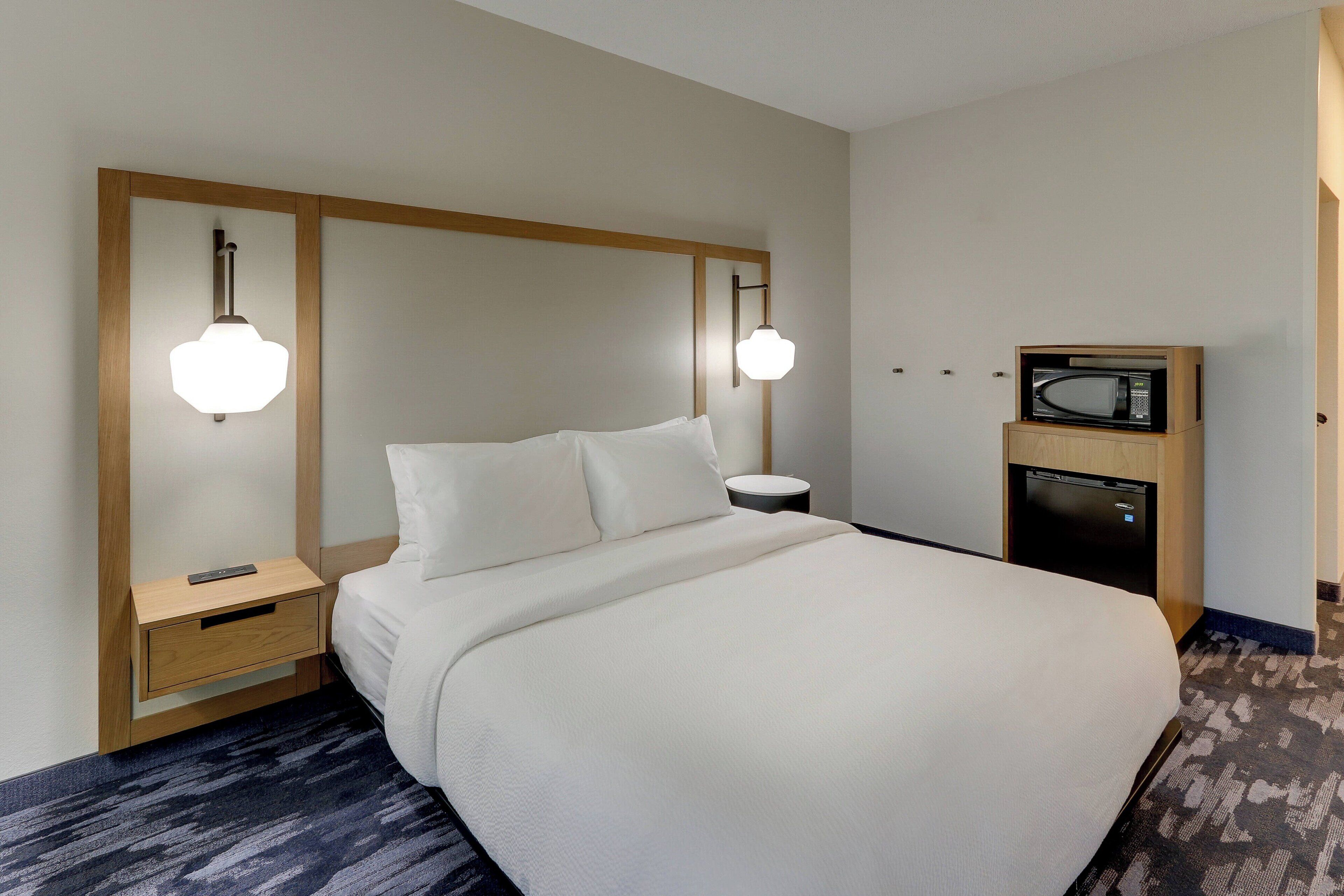 Ein Hotelzimmer mit einem großen Bett, einem Nachttisch mit Lampe, einer Mikrowelle und einer kleinen Schublade.