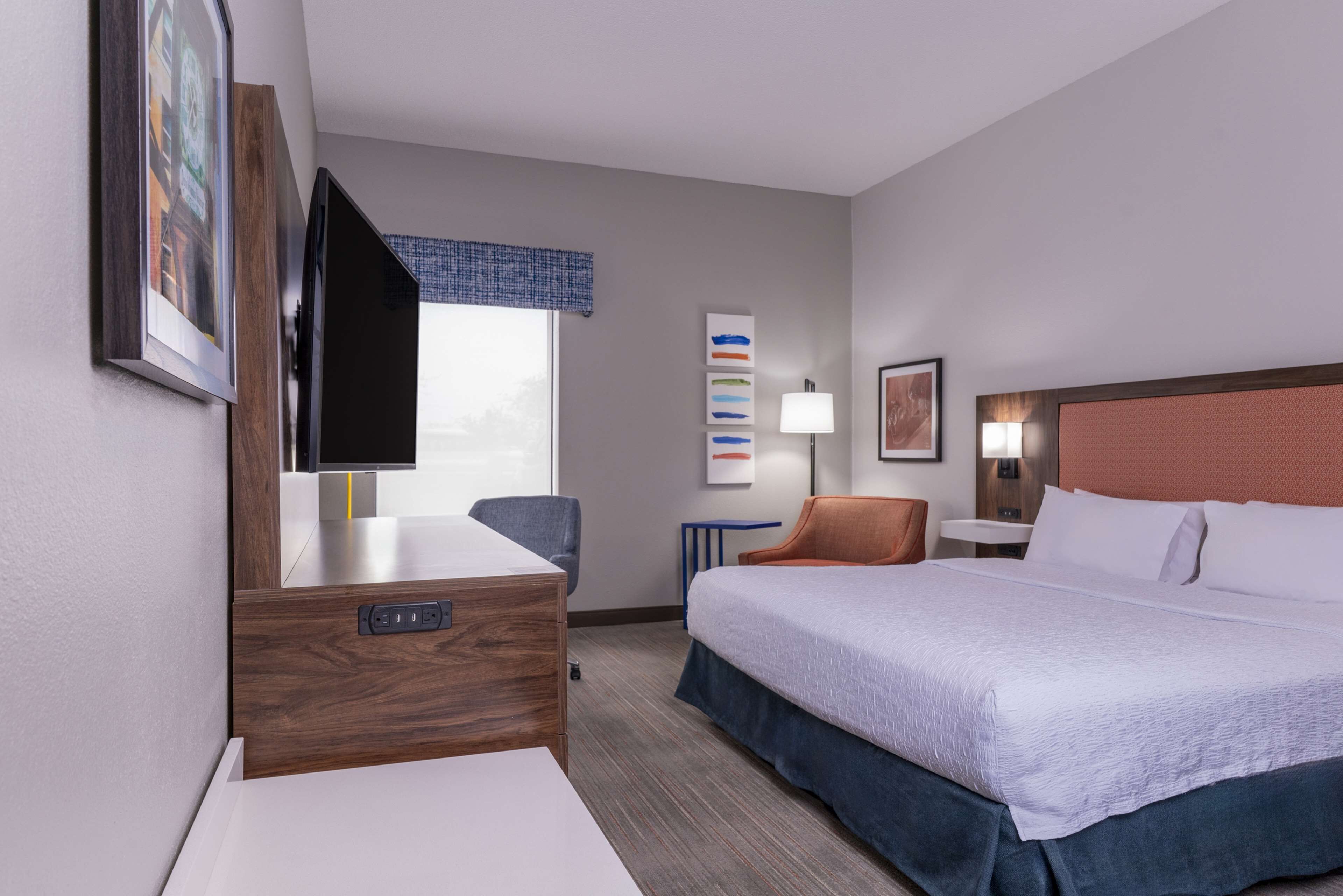 Habitación de hotel con una cama grande, un escritorio con televisor de pantalla plana, una silla, una lámpara y un cuadro en la pared.