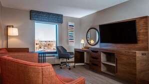 Ein modernes Hotelzimmer mit großem Fenster, einem Schreibtisch mit Flachbildfernseher, einer gemütlichen Sitzecke und einem Wandbild.