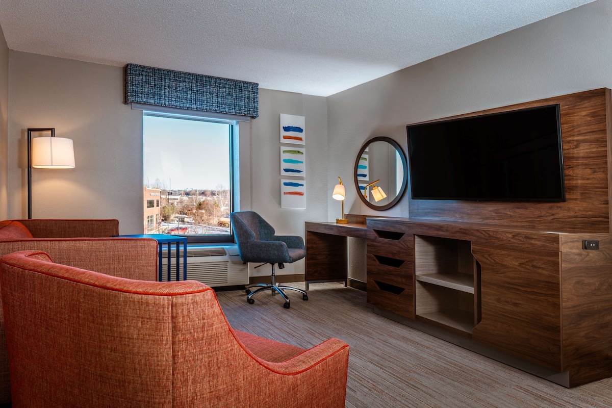 A modern hotel room with a large window, a desk with a flat-screen TV, a comfortable seating area, and a decorative wall piece.