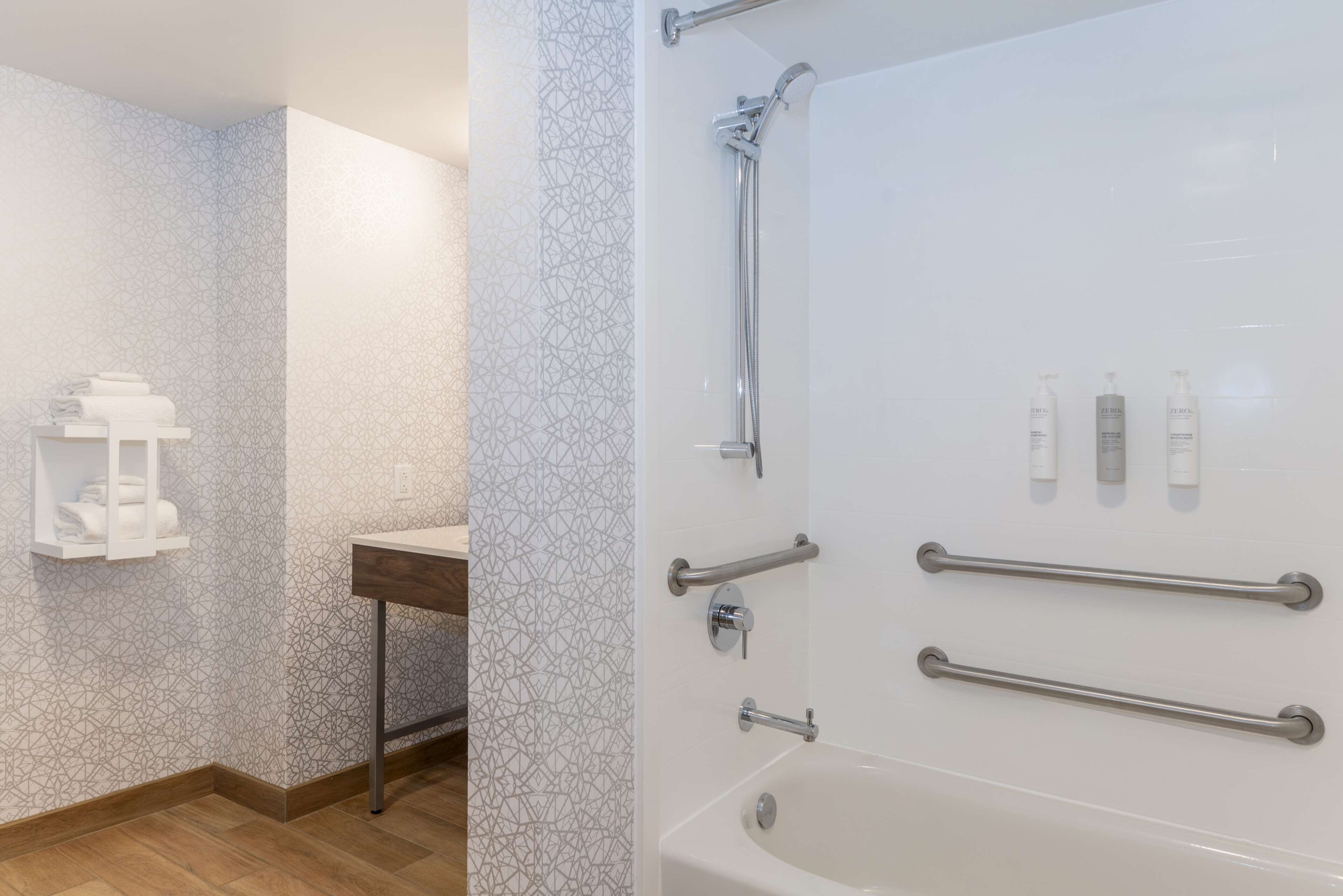 A bathroom with a patterned wall, a sink, a mirror, and a bathtub with grab bars.