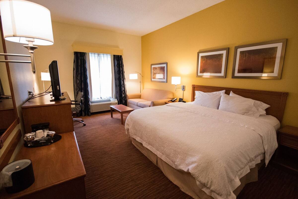 A hotel room with a large bed, a desk with a computer, a chair, a small table, and a window with curtains.