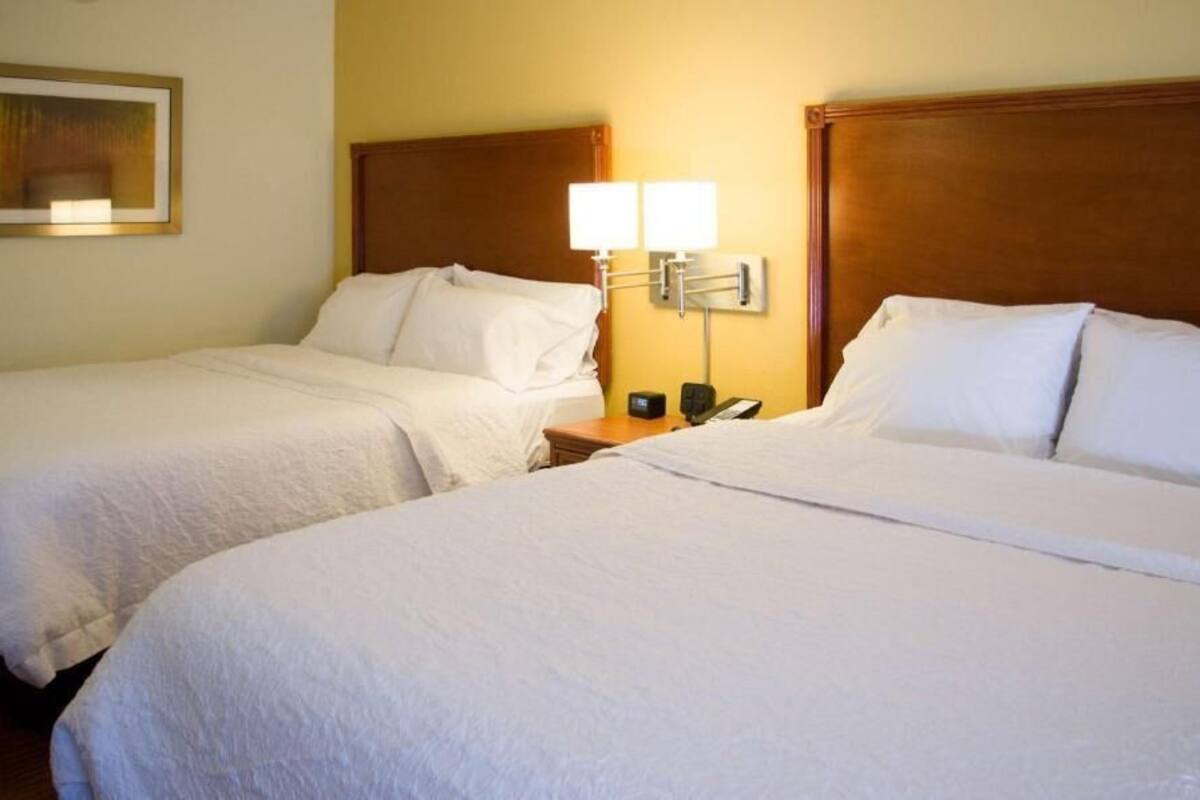 A hotel room with two single beds, a nightstand with a lamp, and a framed picture on the wall.