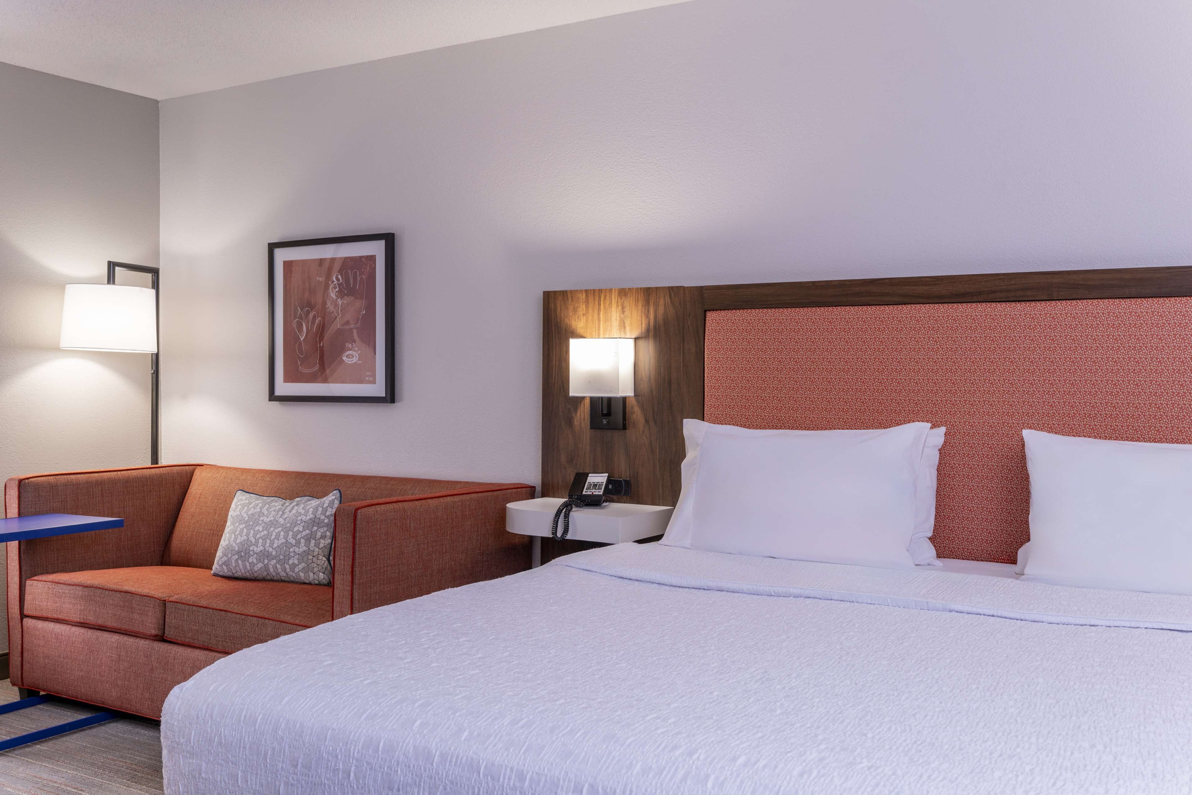 A hotel room with a large bed, a sofa, a bedside table with a lamp, and a framed picture on the wall.