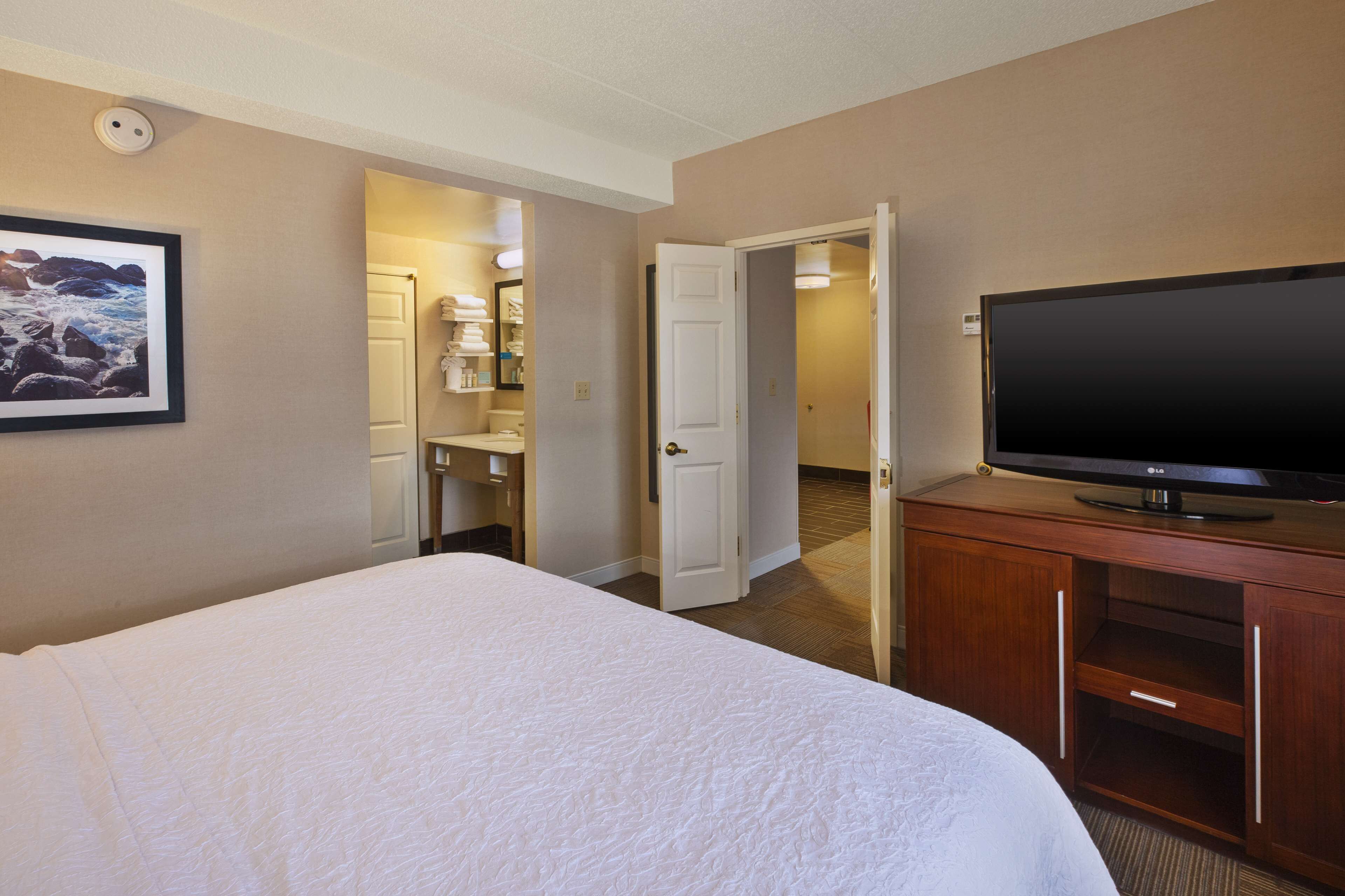 Une chambre d’hôtel avec un grand lit, un téléviseur posé sur un meuble en bois, une salle de bain avec un lavabo et un miroir, et un couloir menant à une autre chambre.