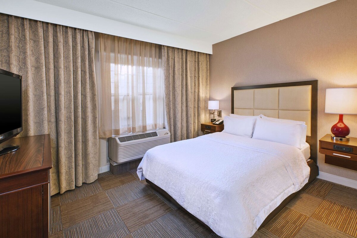 A hotel room with a large bed, a TV, an air conditioning unit, a bedside lamp, and a nightstand.