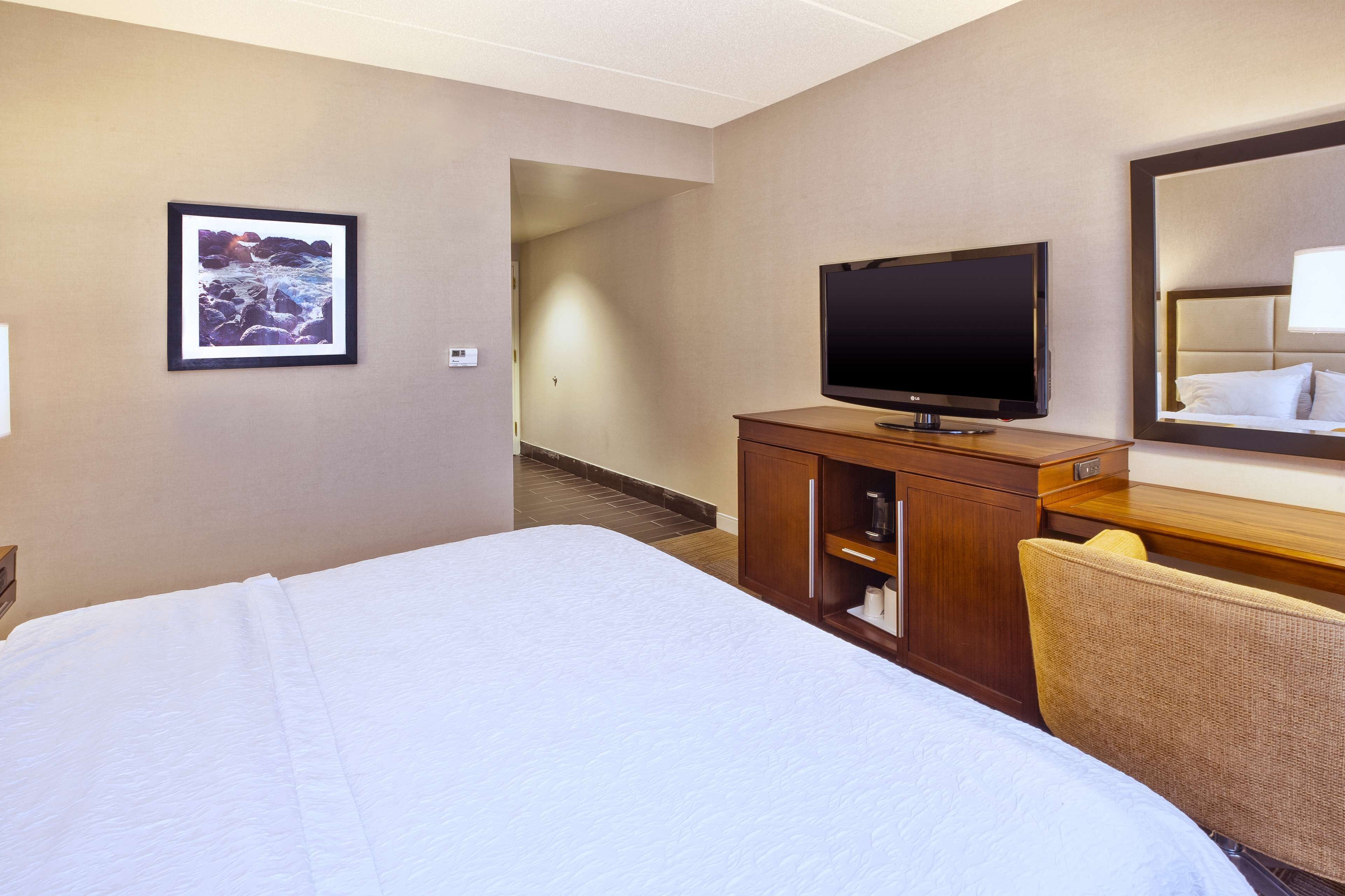 Una habitación de hotel con una cama grande, televisión, cómoda de madera, una silla y un cuadro en la pared.