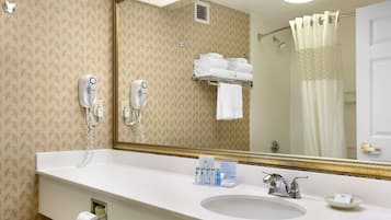 Un baño de hotel con un espejo grande, un lavabo con artículos de aseo, un perchero y una zona de ducha.