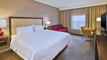 A hotel room with a large bed, a desk with a television, a red chair, and a desk lamp.