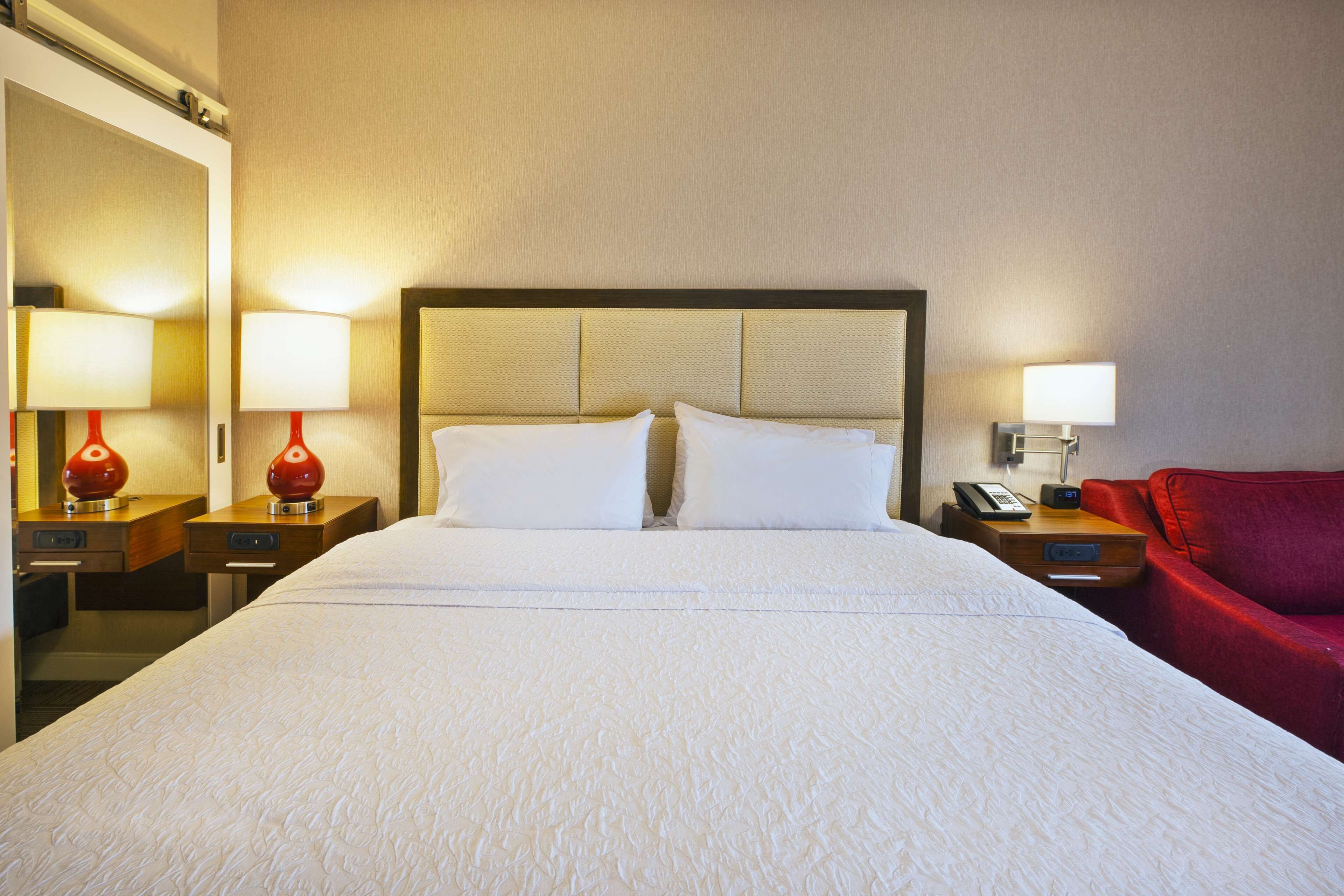 Una habitación de hotel con una cama grande, dos mesitas de noche con lámparas, una silla roja y un teléfono en la pared.
