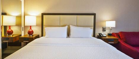Una habitación de hotel con una cama grande, dos mesitas de noche con lámparas, una silla roja y un teléfono en la pared.