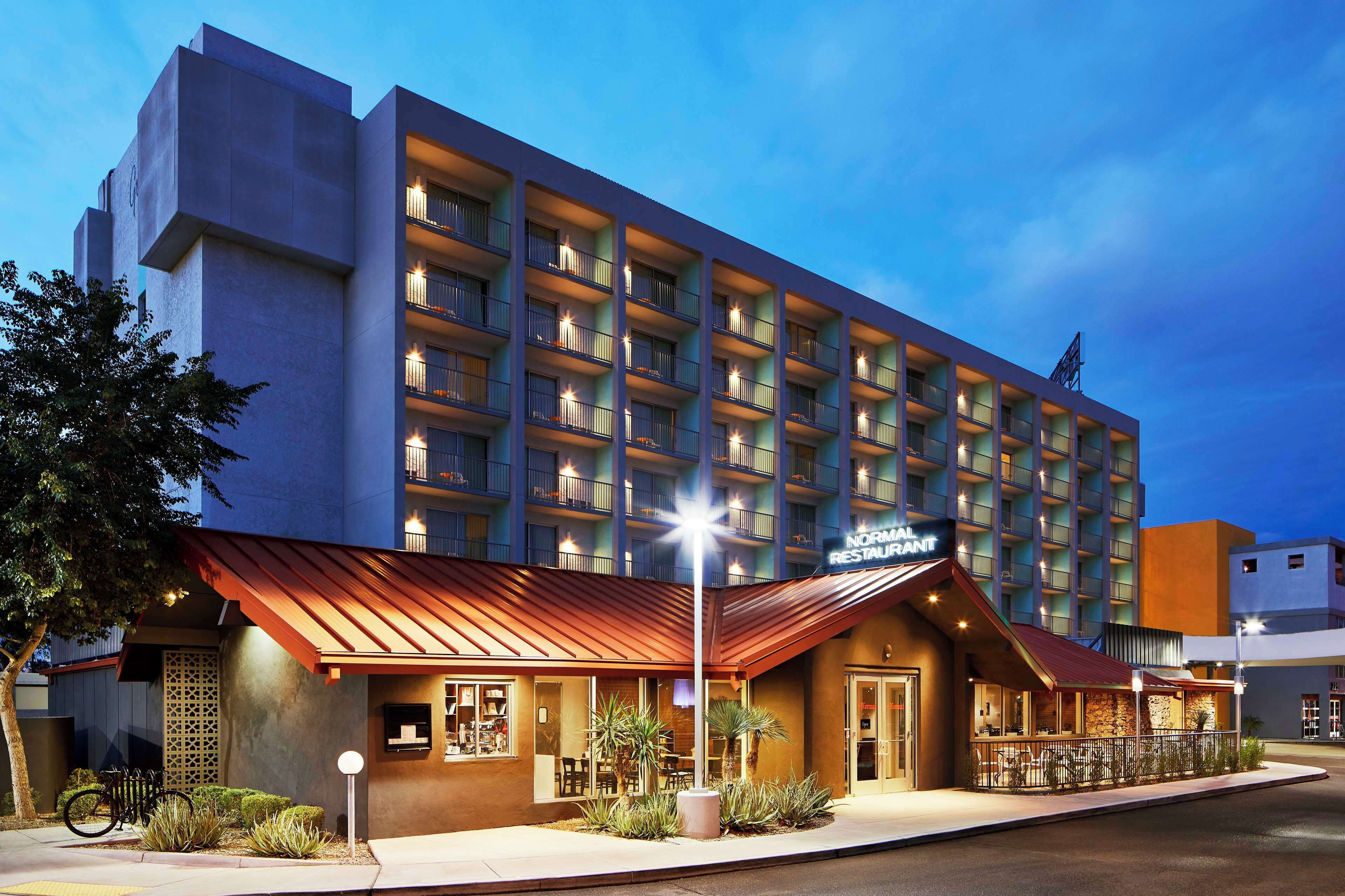 Um hotel de vários andares com design moderno, com varandas em cada andar, um restaurante com área externa e uma entrada bem iluminada.