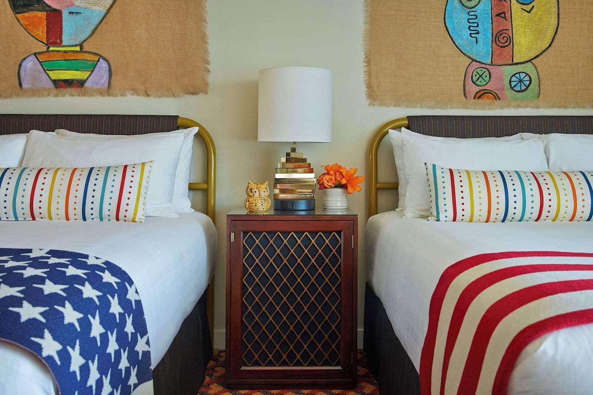 A hotel room with two beds, each with a striped pillow and a star-patterned blanket. A small table with a lamp and a decorative item is between the beds.