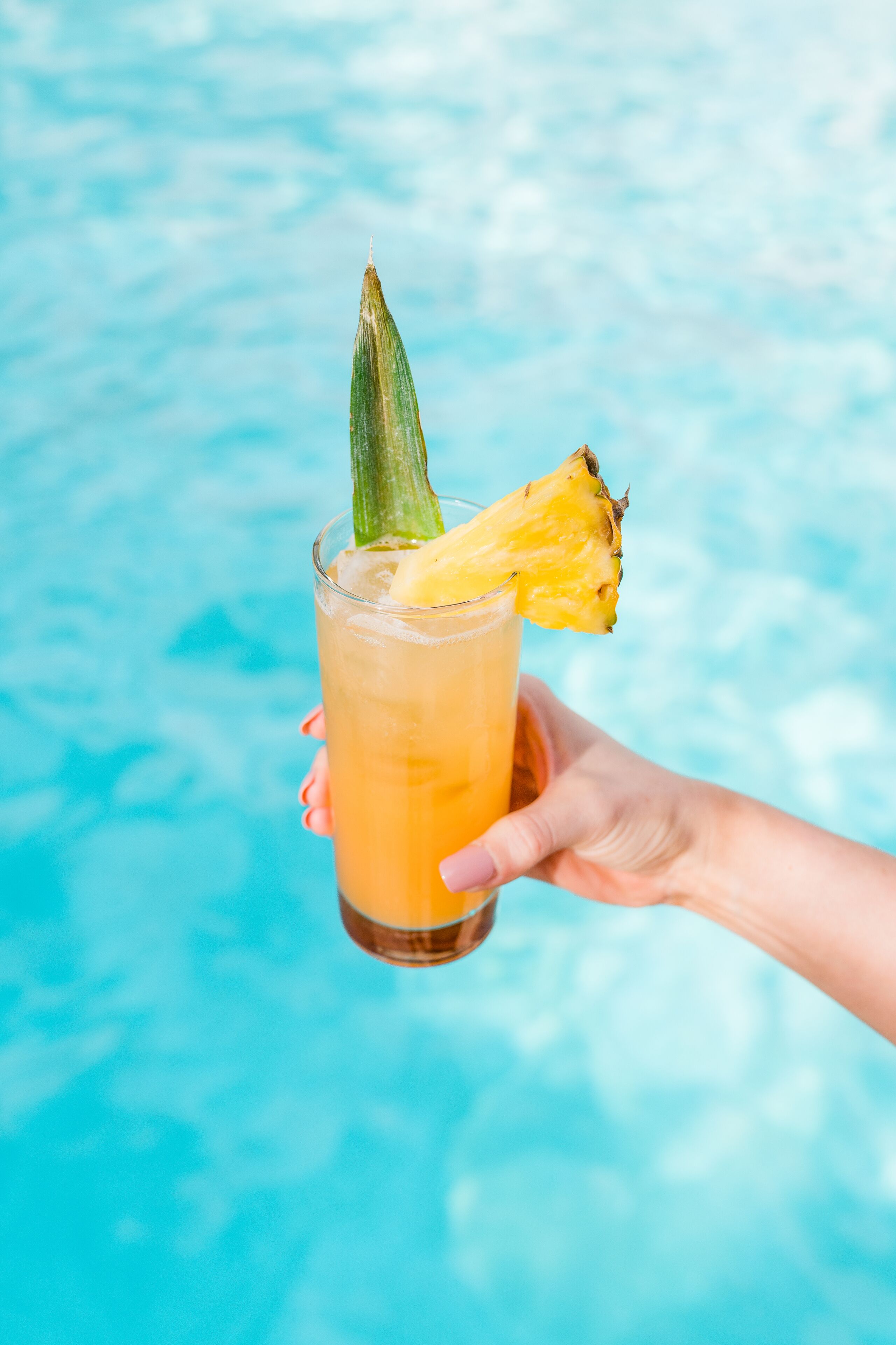 Una mano sosteniendo un vaso con una decoración de piña, con una piscina al fondo.