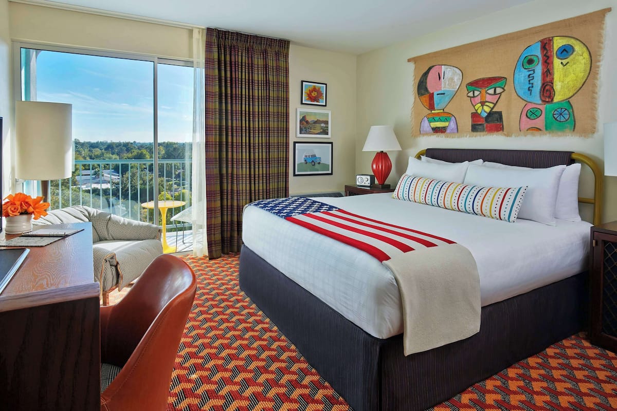 A hotel room with a large bed, a desk, a chair, and a balcony with a view of the city.