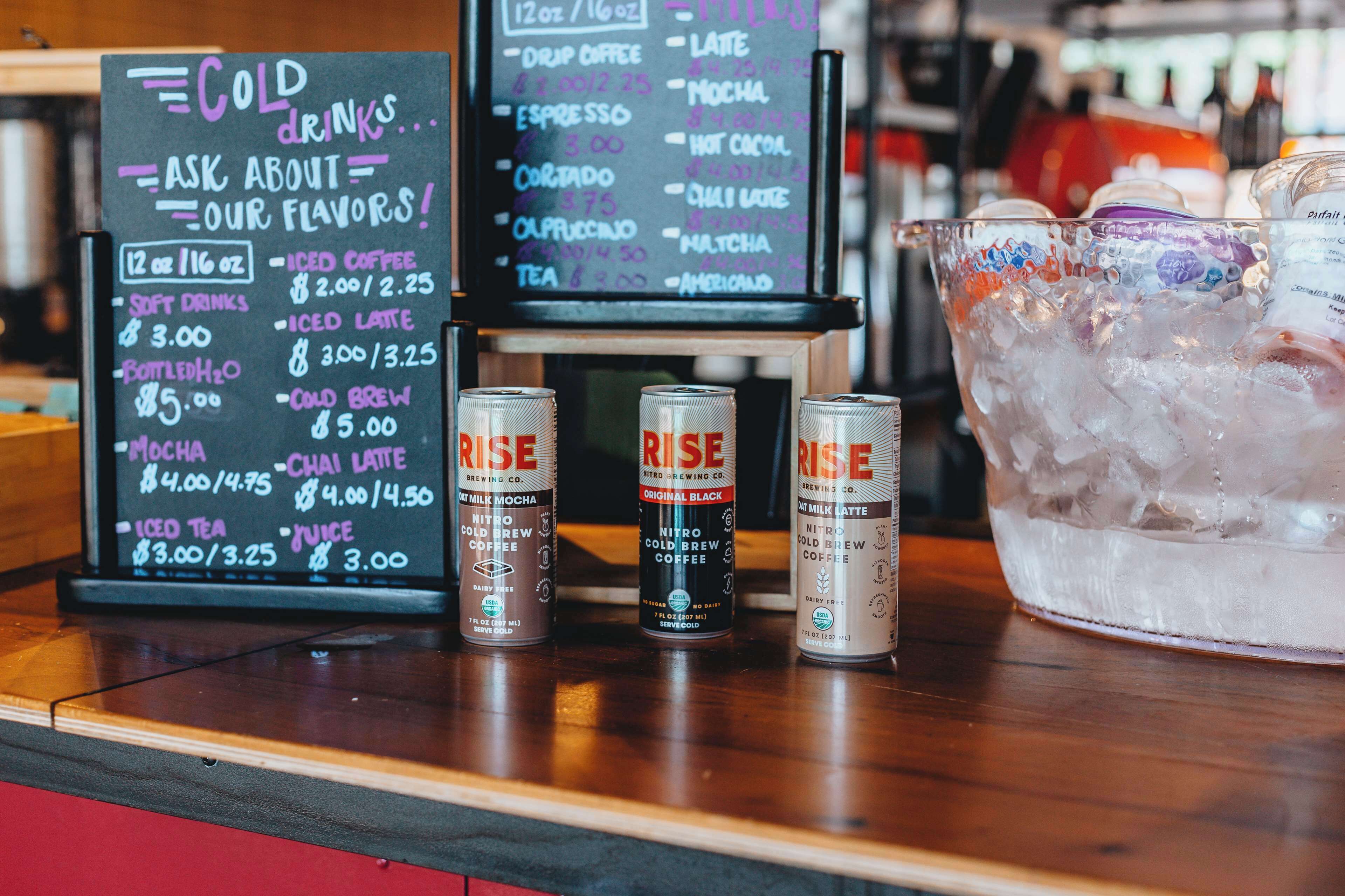A coffee shop counter with a menu for cold drinks and a selection of Rise brand coffee cans.