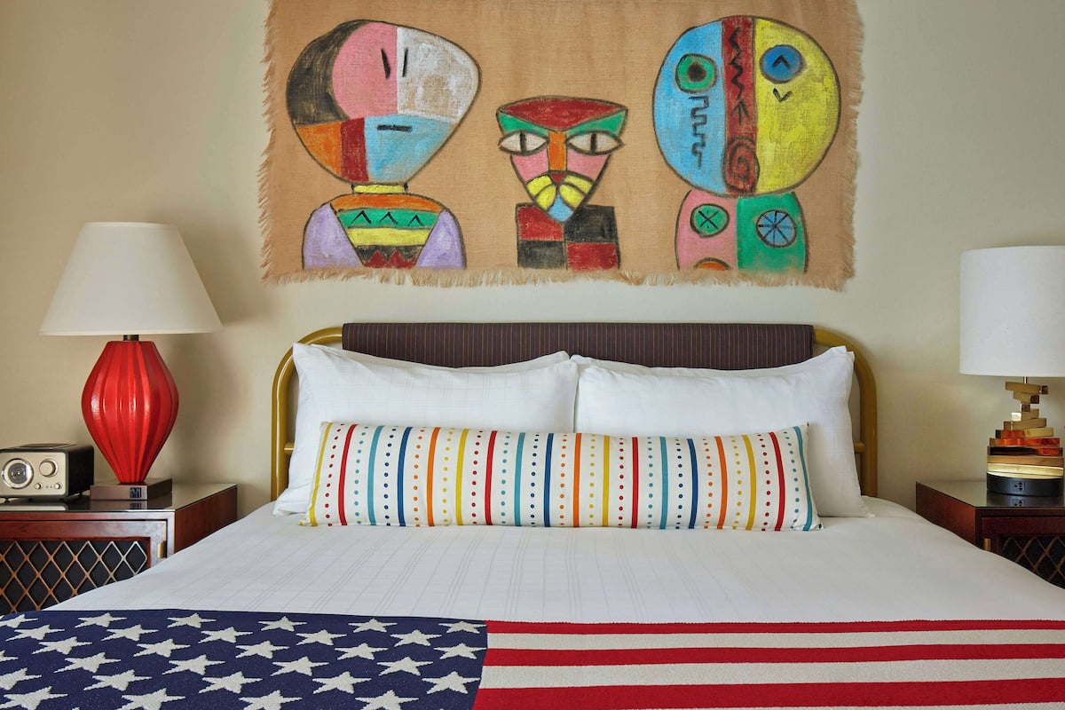 A hotel room with a bed, two bedside lamps, a radio, and a wall painting.