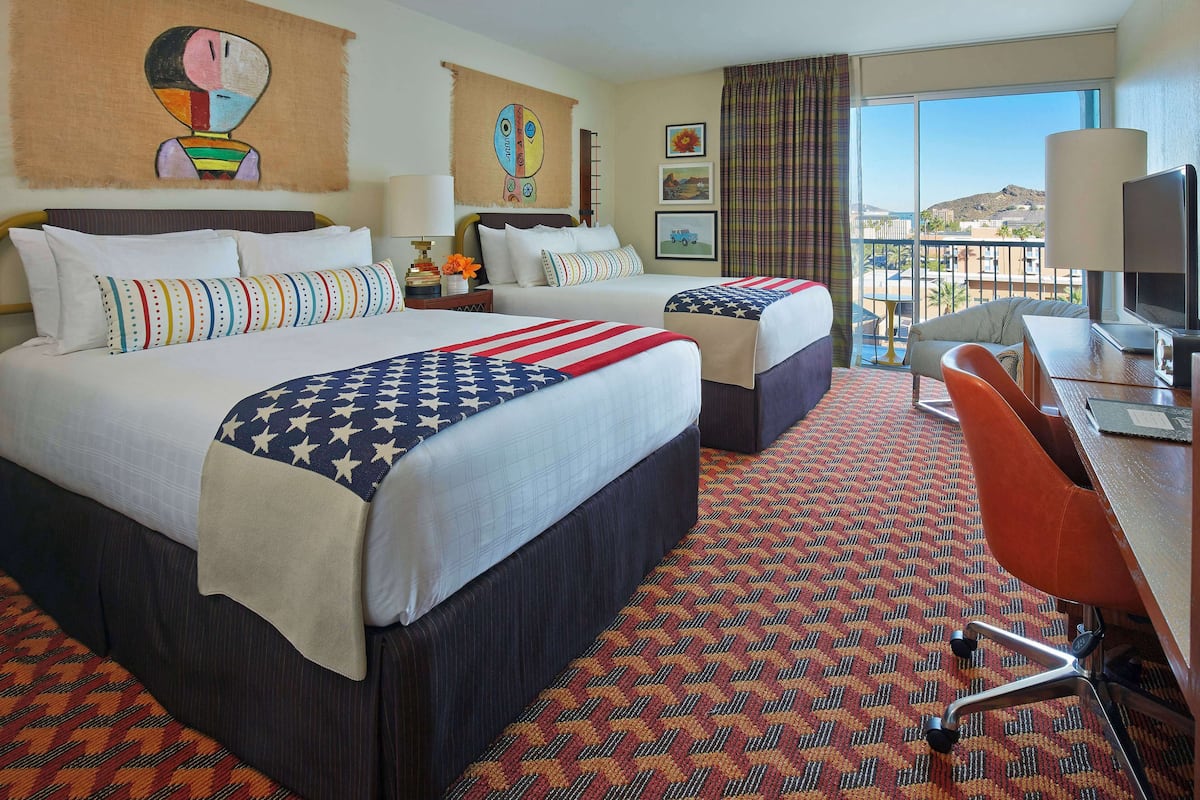 A hotel room with two beds, a desk with a chair, and a large window offering a view of the landscape.