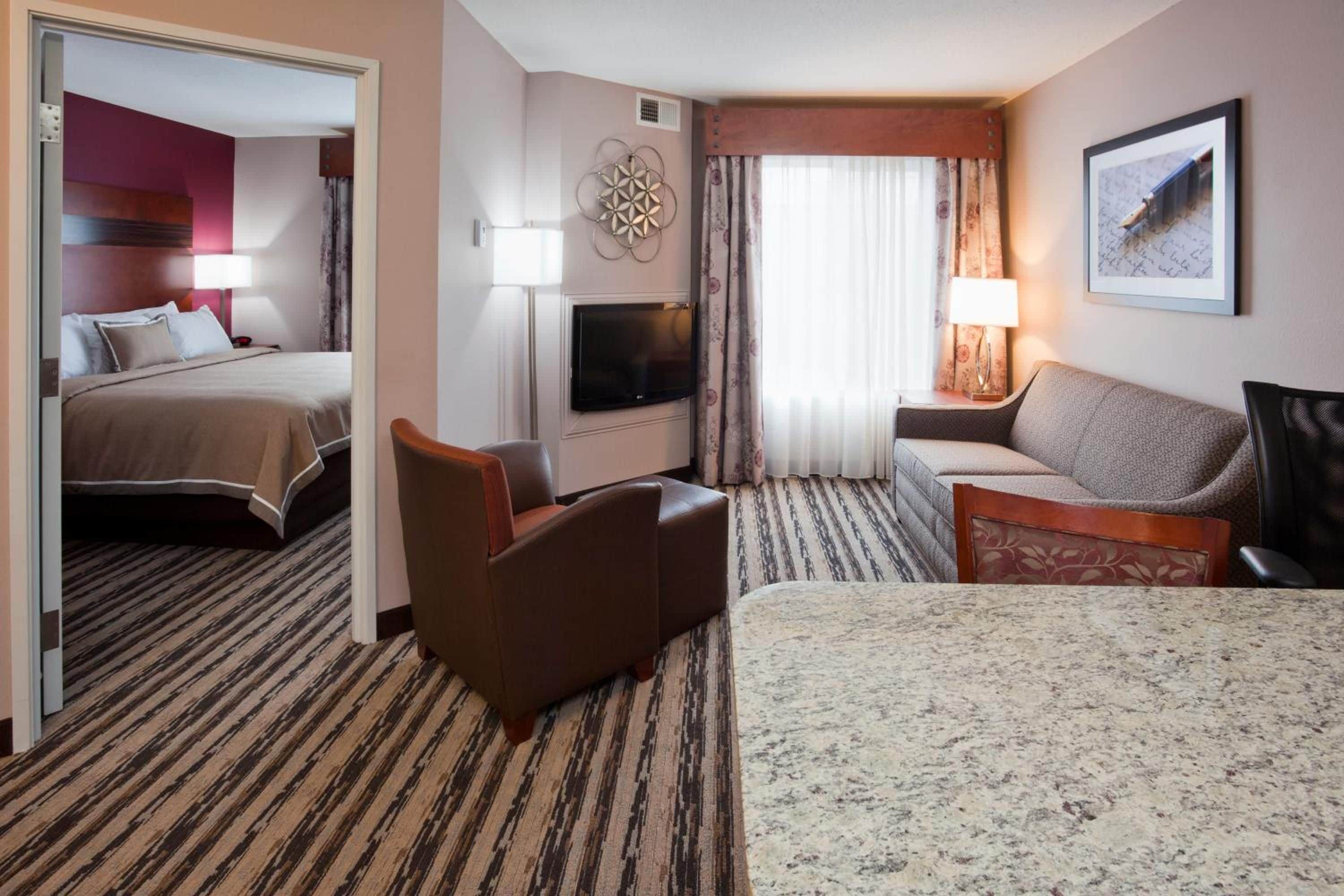 Ett hotellrum med en säng, en soffa, en stol, en tv och ett fönster med gardiner.