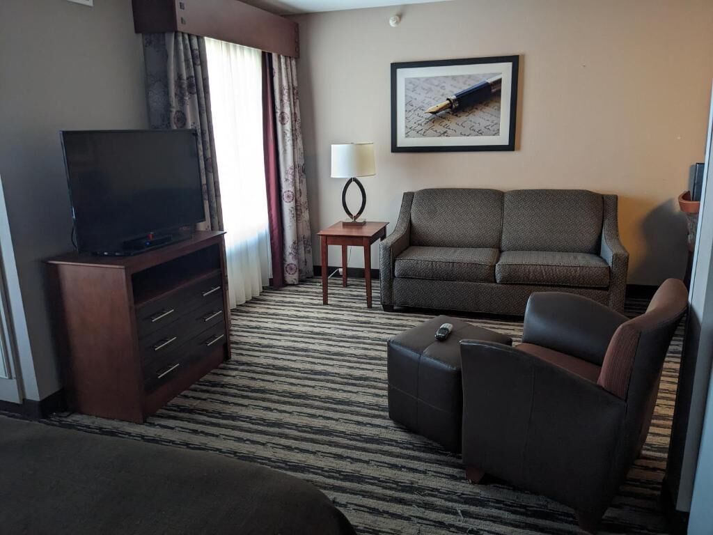 Een hotelkamer met een flatscreen televisie op een houten standaard, een bank, een kleine tafel met een lamp en een ingelijst kunstwerk aan de muur.