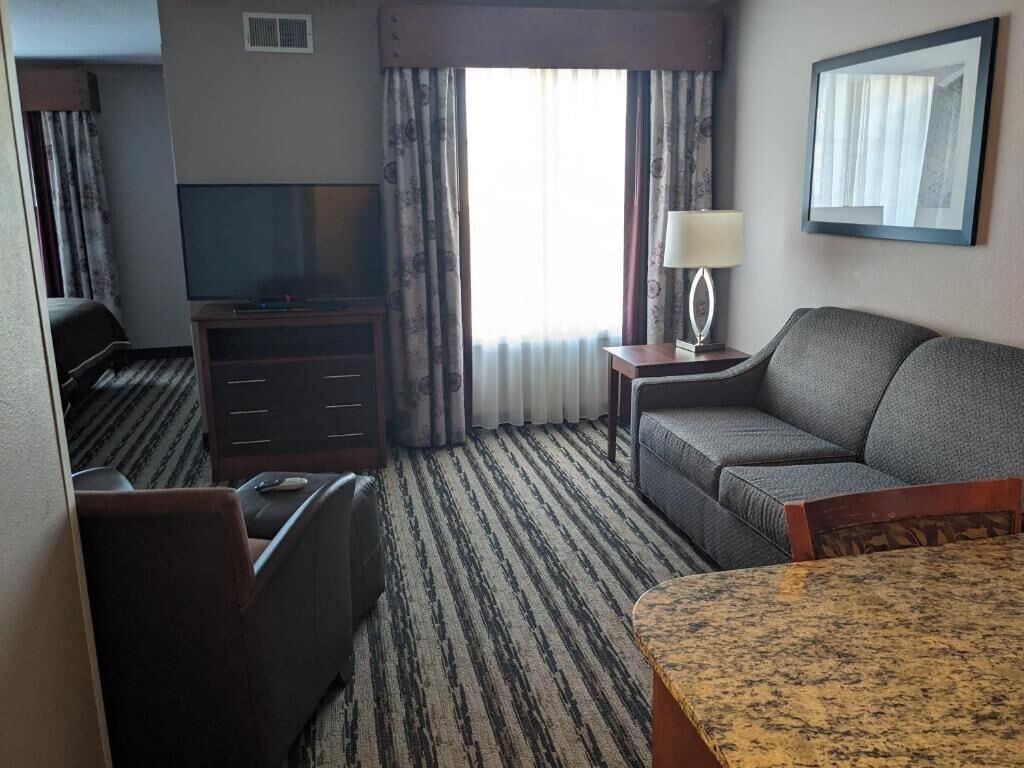 Habitación de hotel con televisión, sofá, escritorio, lámpara y ventana con cortinas.