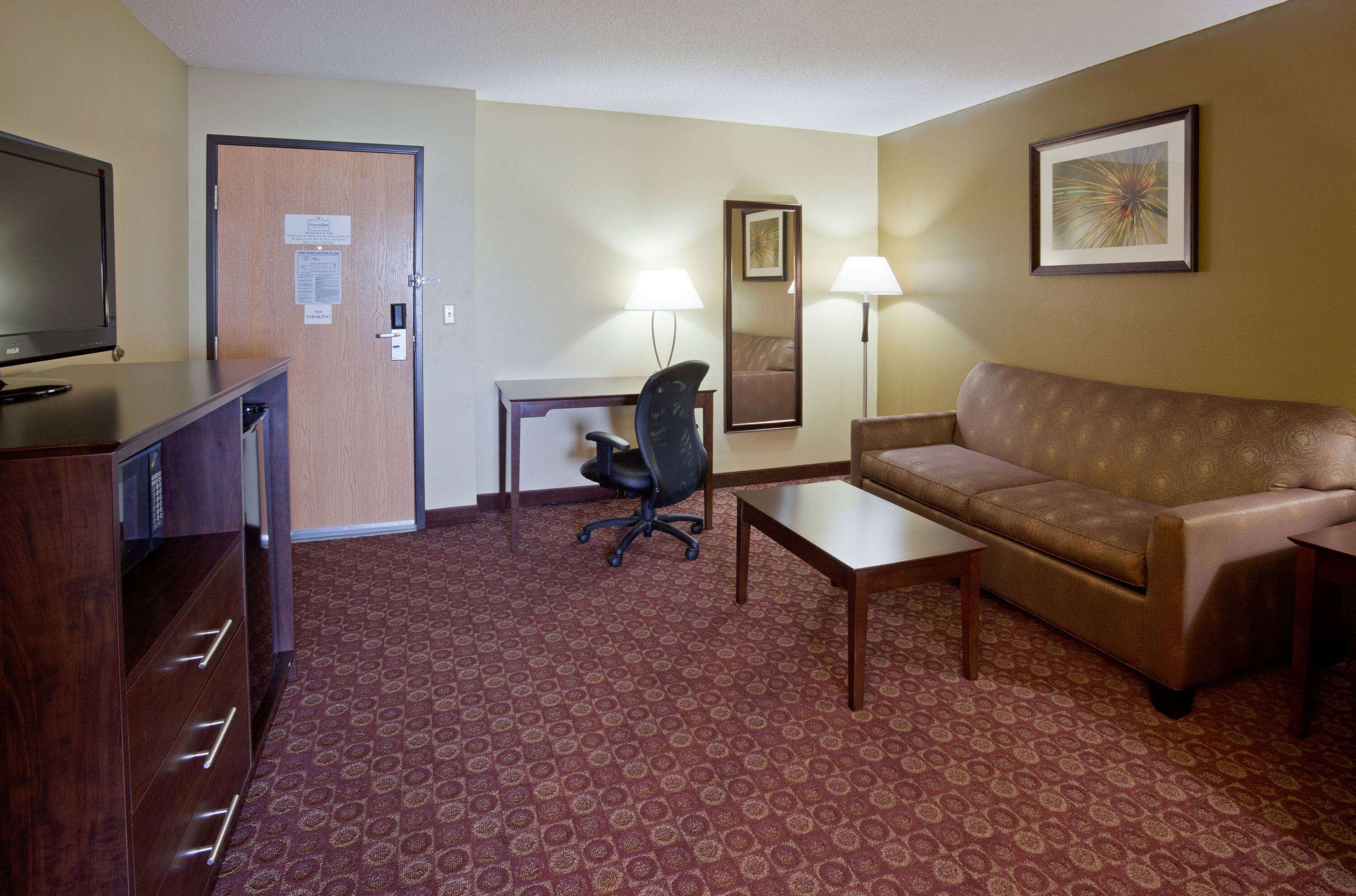 A hotel room with a brown leather sofa, a wooden coffee table, a desk with a chair, a television on a wooden cabinet, and a patterned carpet.