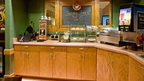 Un comptoir de café avec un tableau noir indiquant la date et la température, un appareil à gaufres, une machine à café, une vitrine remplie de pâtisseries et un distributeur de jus.