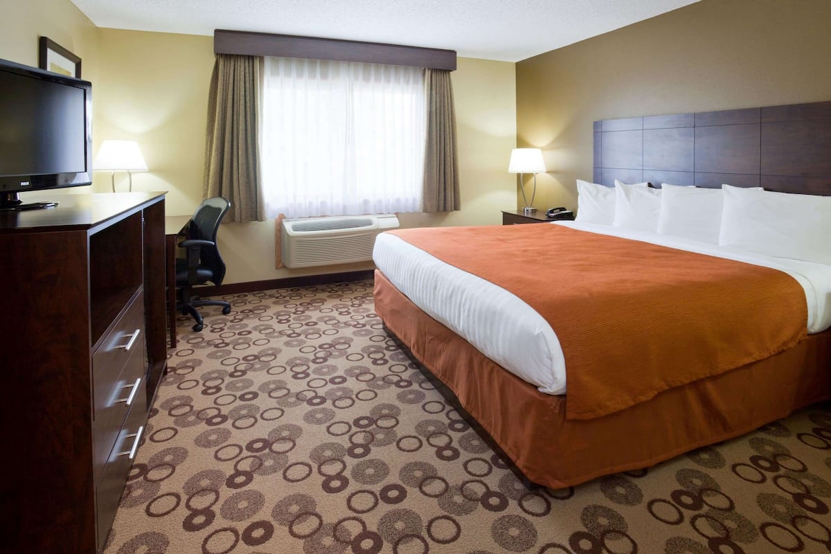 A hotel room with a large bed, a desk with a television, a chair, an air conditioning unit, and a window with curtains.