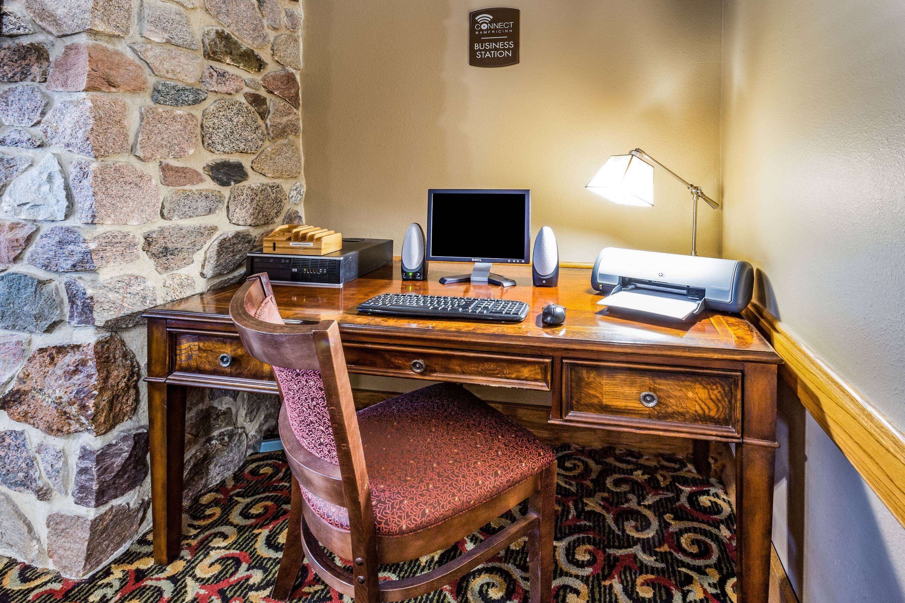 A wooden desk with a computer, keyboard, mouse, and speakers. There is a chair with a patterned cushion. A sign on the wall reads "CONNECT KAMIBIBIDON BUSINESS STATION."