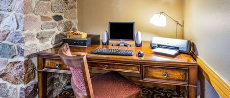 A wooden desk with a computer, keyboard, mouse, and speakers. There is a chair with a patterned cushion. A sign on the wall reads "CONNECT KAMIBIBIDON BUSINESS STATION."