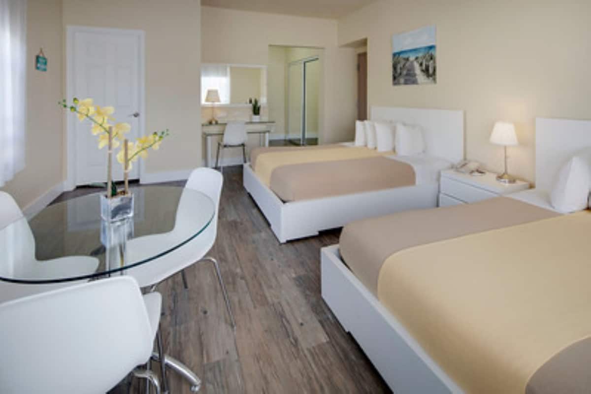A hotel room with two beds, a desk with a chair, a glass table, and a small vase with flowers.