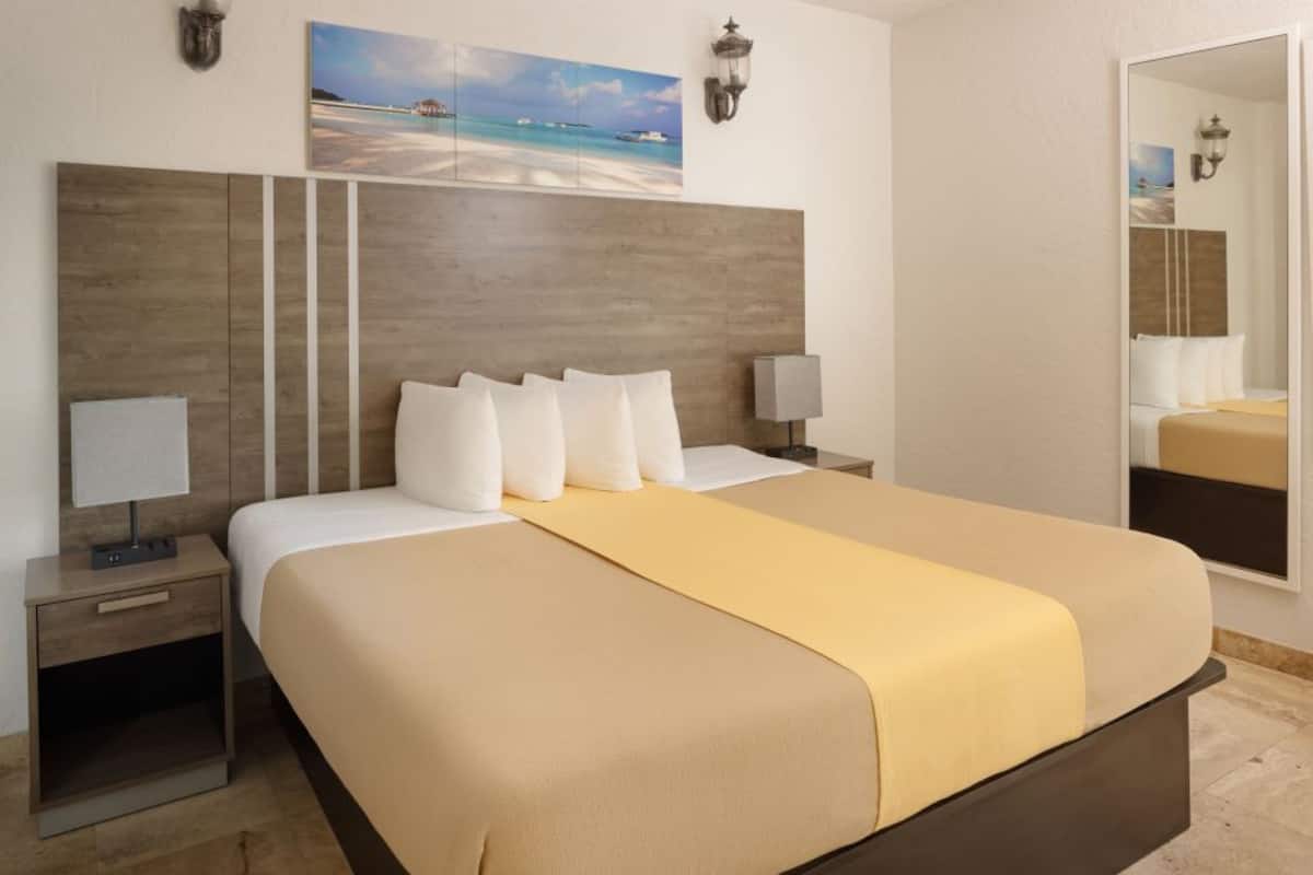 A hotel room with a large bed, two bedside tables, a nightstand, a wall-mounted lamp, and a painting of a beach scene.