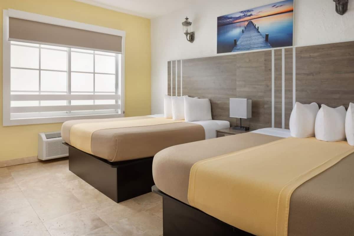 A hotel room with two beds, a large window with blinds, a wall-mounted artwork, and a nightstand with a lamp.