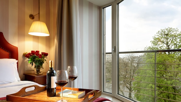 A hotel room with a bed, a bedside table with a vase of red roses, a tray with a bottle of wine and two glasses, and a window with a view of trees. - Eurostars Montgomery (Brussels)