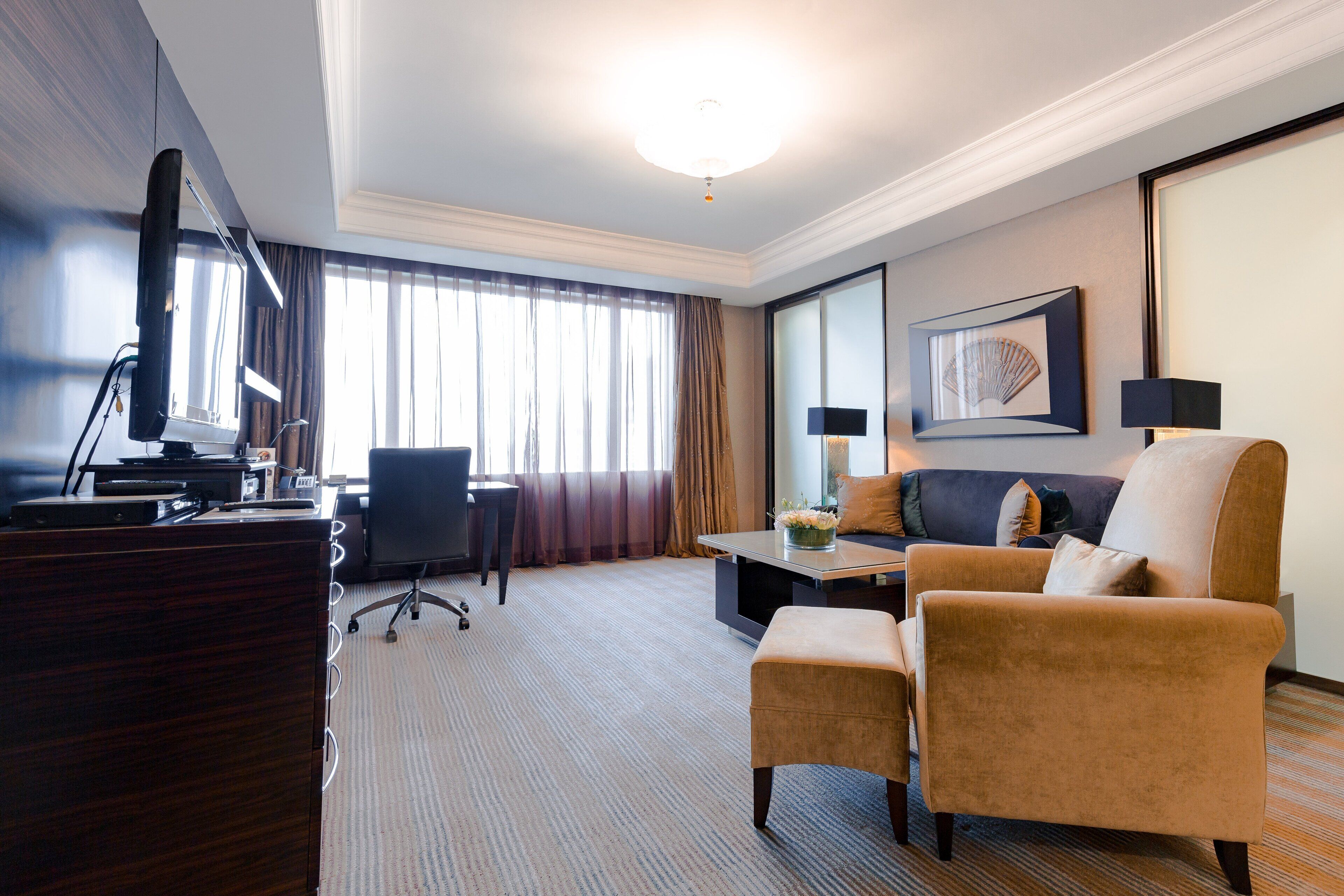 A hotel room with a desk, a sofa, an ottoman, a TV, and a large window with curtains.