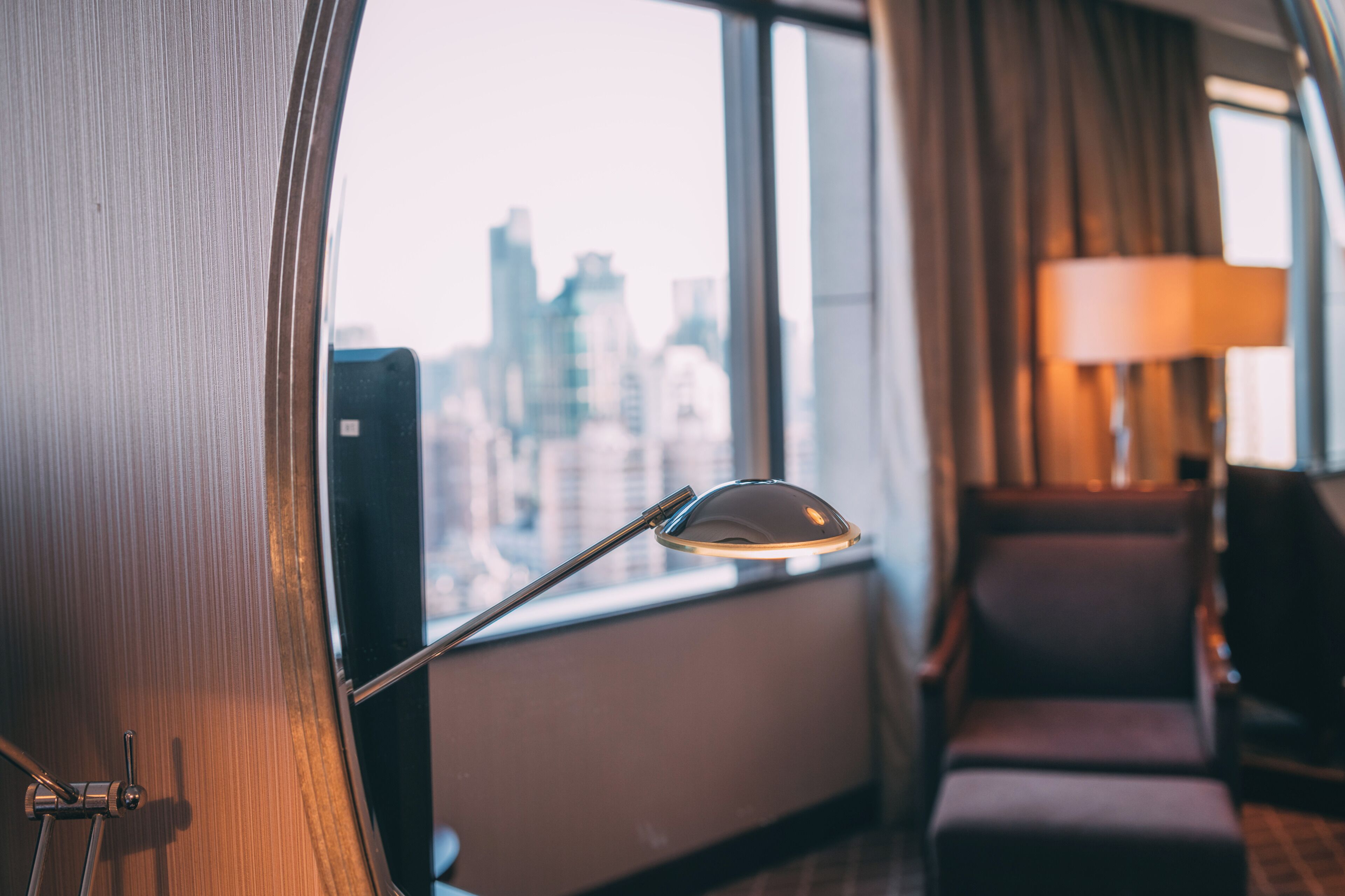 Una habitación con vista a un paisaje urbano a través de una ventana, una lámpara y un sillón.