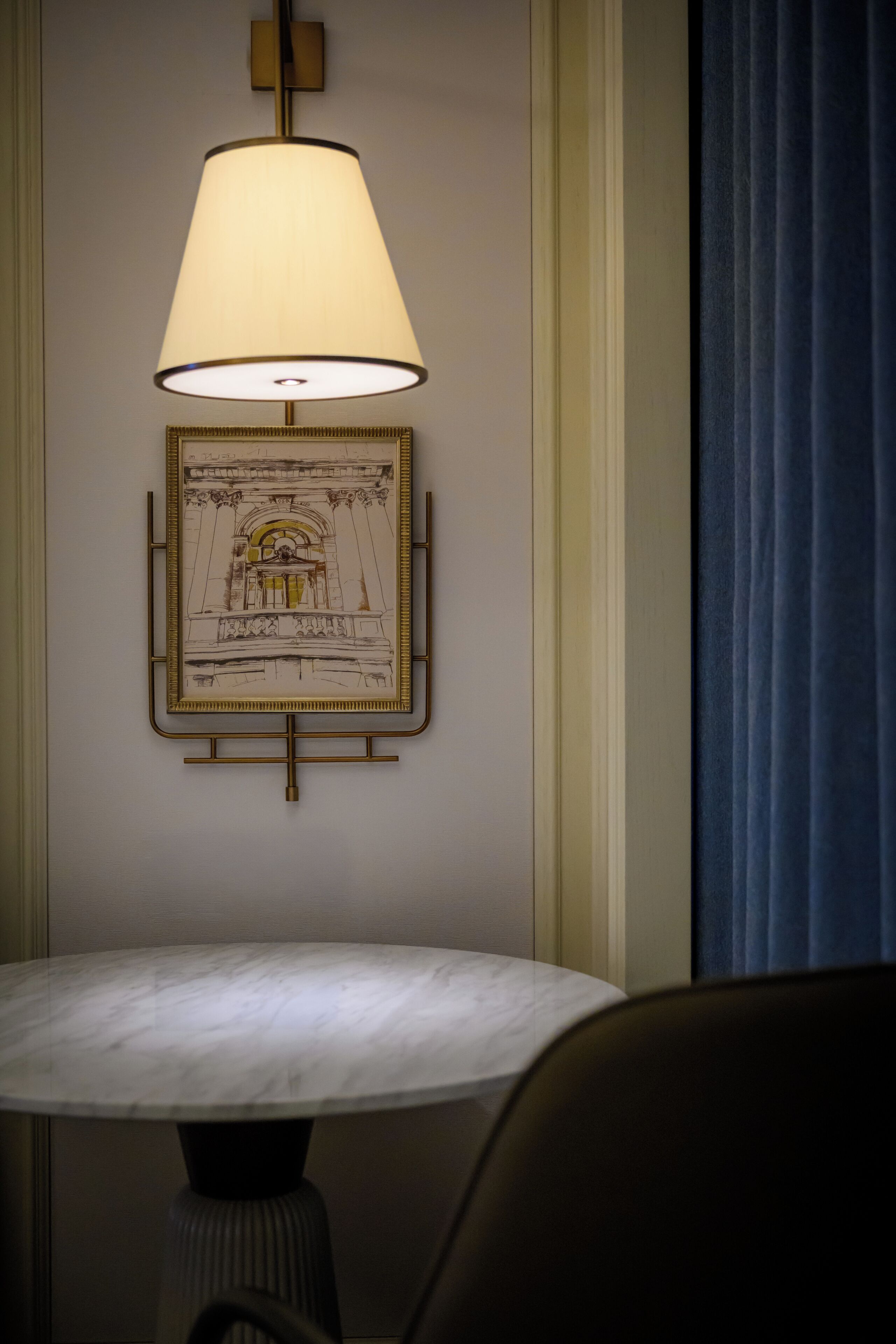 Una lampada a parete con un ampio paralume, un disegno architettonico incorniciato e un tavolo tondo con una sedia.