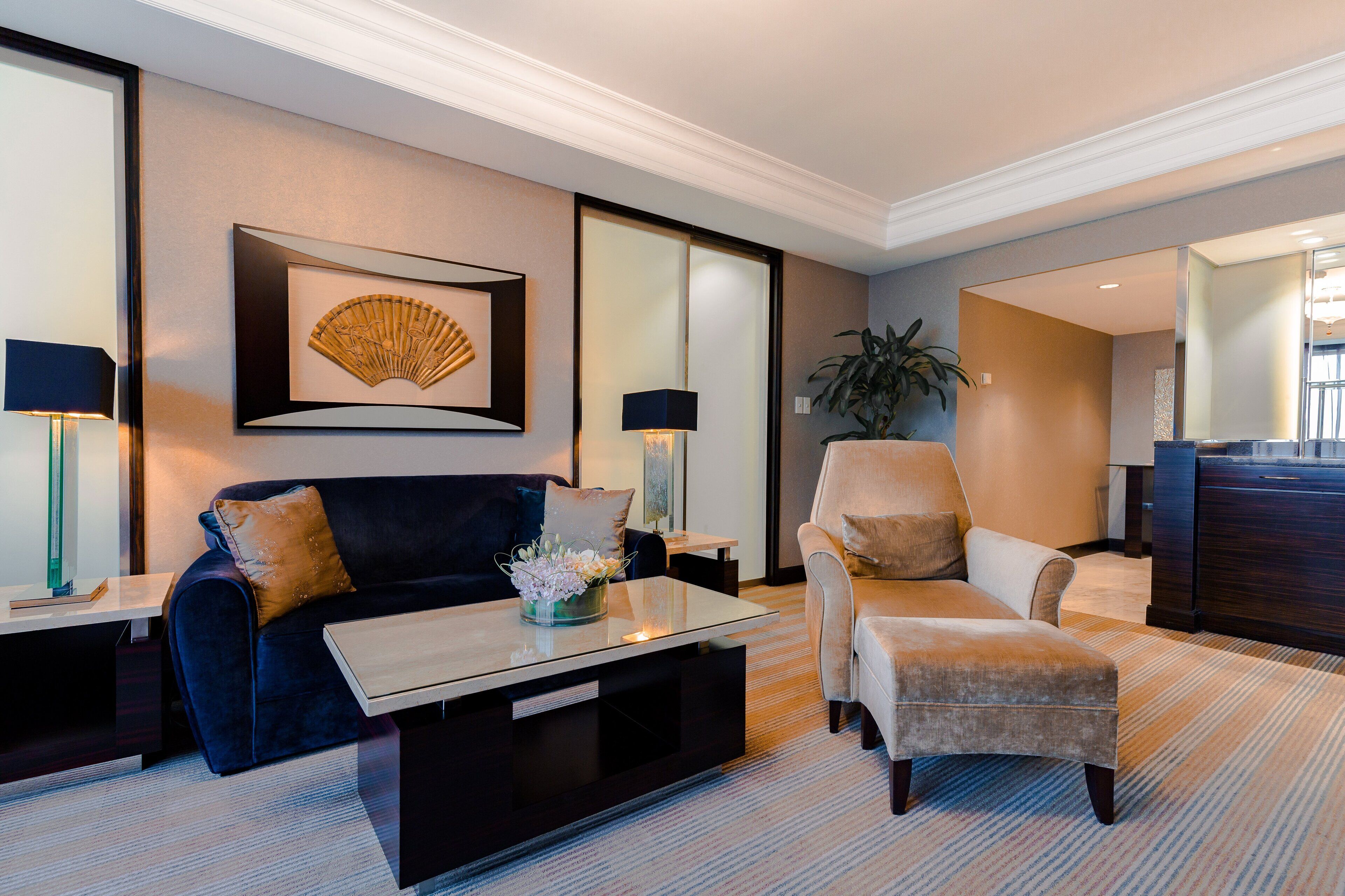 A modern living room with a dark blue sofa, a beige armchair, a glass coffee table, and a large framed artwork on the wall.
