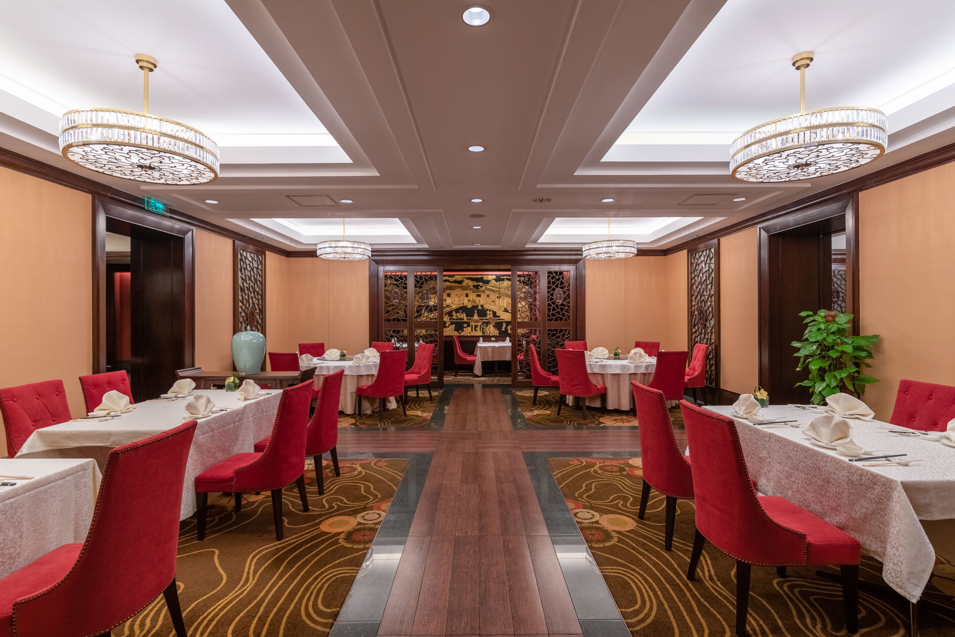 Um espaço de refeições de hotel com mesas preparadas para o serviço, cadeiras estofadas vermelhas e lustres.
