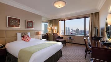 Ein Hotelzimmer mit einem großen Bett, einem Schreibtisch mit Spiegel, einem Sessel und Blick auf die Stadtlandschaft.