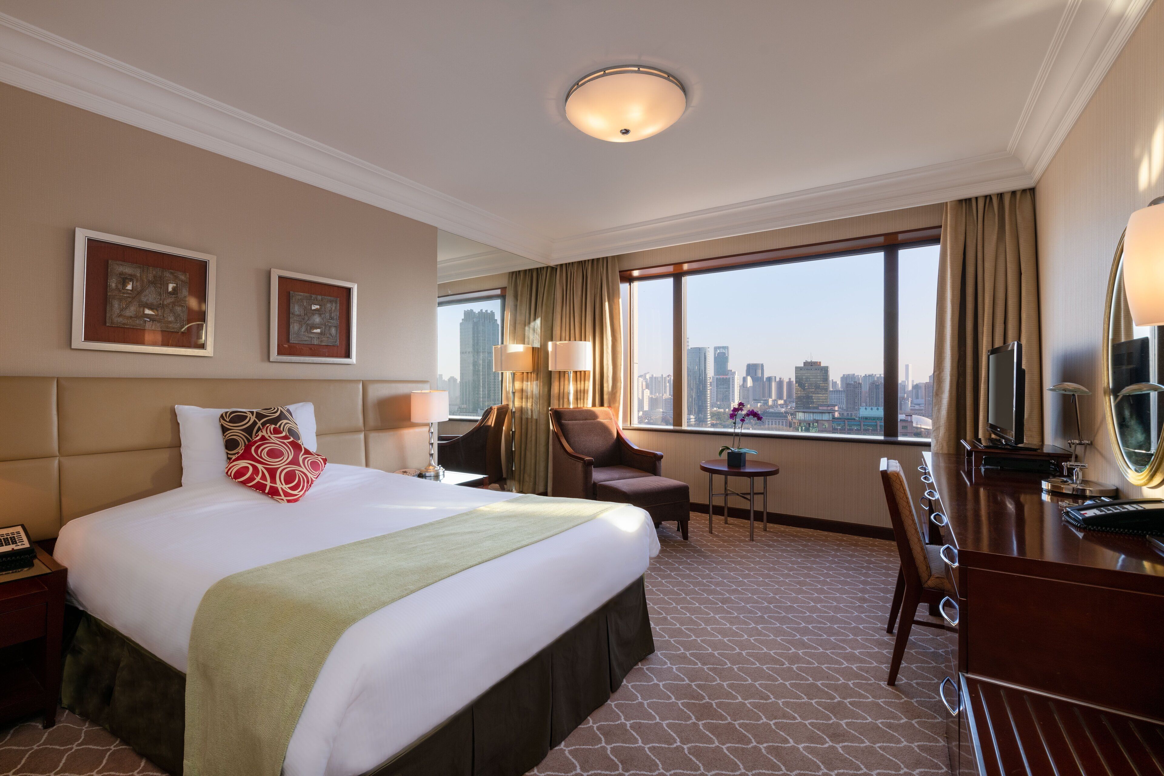 A hotel room with a large bed, a desk with a mirror, a chair, a small table, and a view of the city skyline.