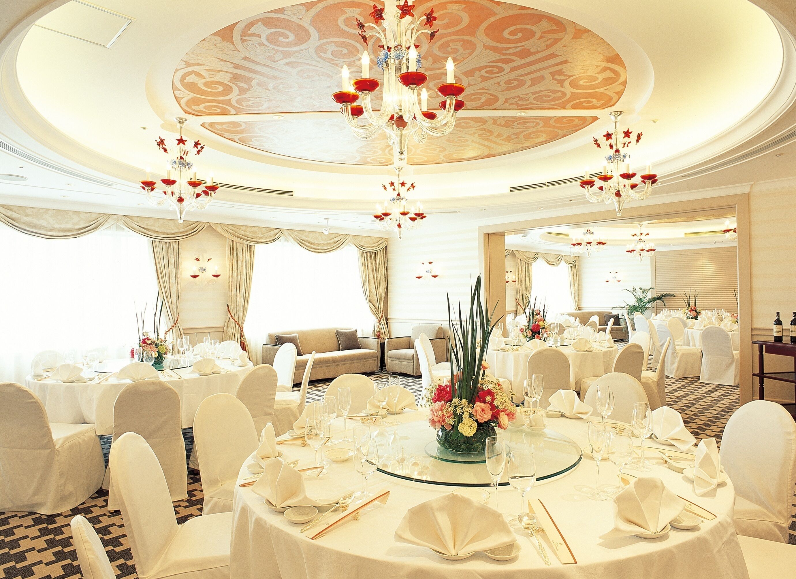 A banquet hall with round tables, white chairs, chandeliers, and a decorative ceiling.
