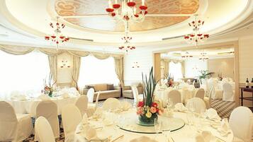 A banquet hall with round tables, white chairs, chandeliers, and a decorative ceiling.