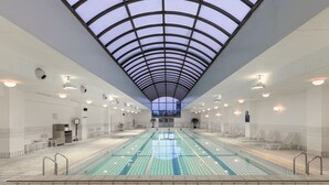 Una piscina interior con techos altos y tragaluces, rodeada de sillas de descanso y un suelo de baldosas.