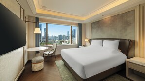 Habitación de hotel con una cama grande, un escritorio, una silla, un pequeño reposapiés, un televisor y vista a la ciudad.
