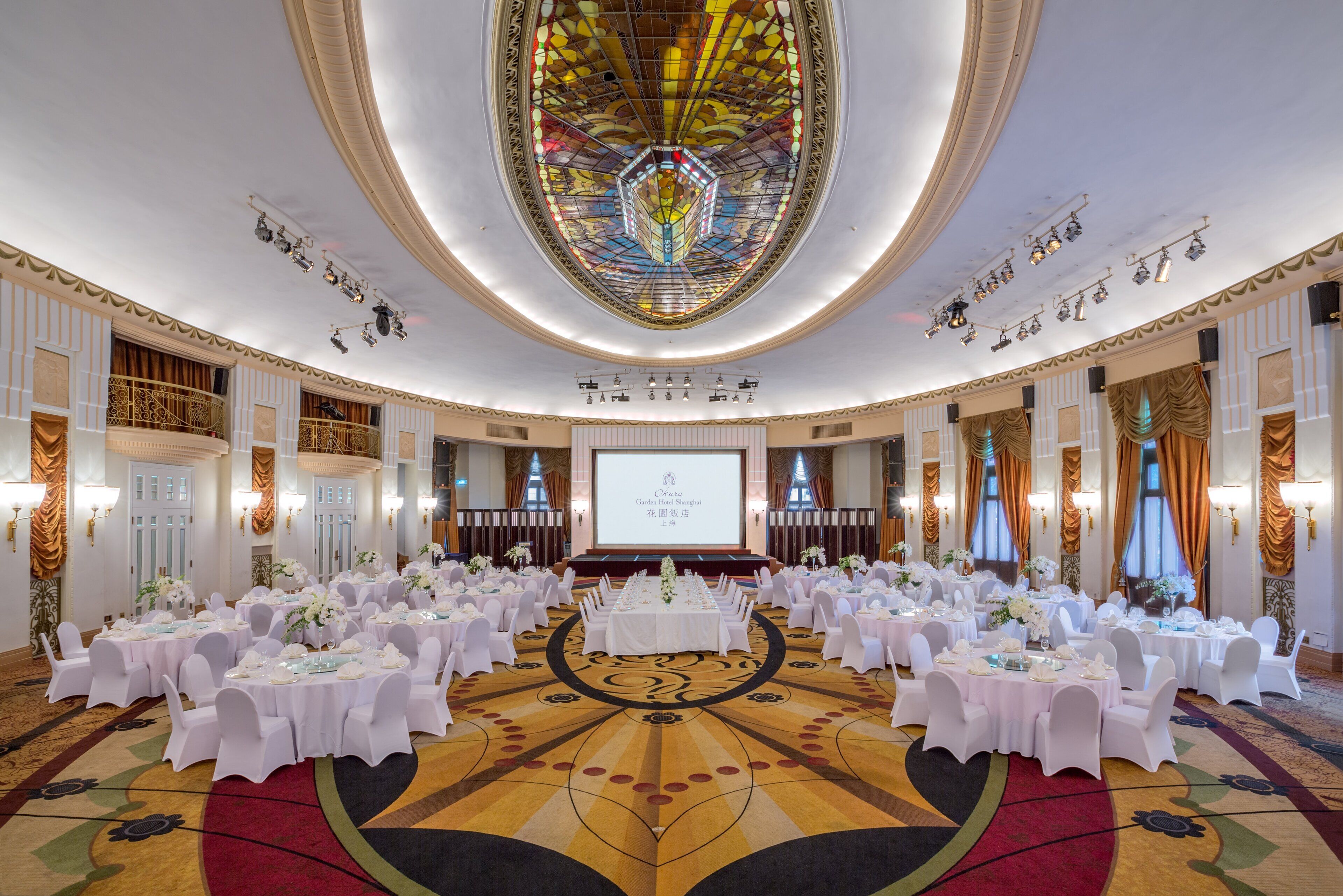 A large banquet hall with round tables set for a formal event, a large screen at the front, and ornate ceiling decorations.