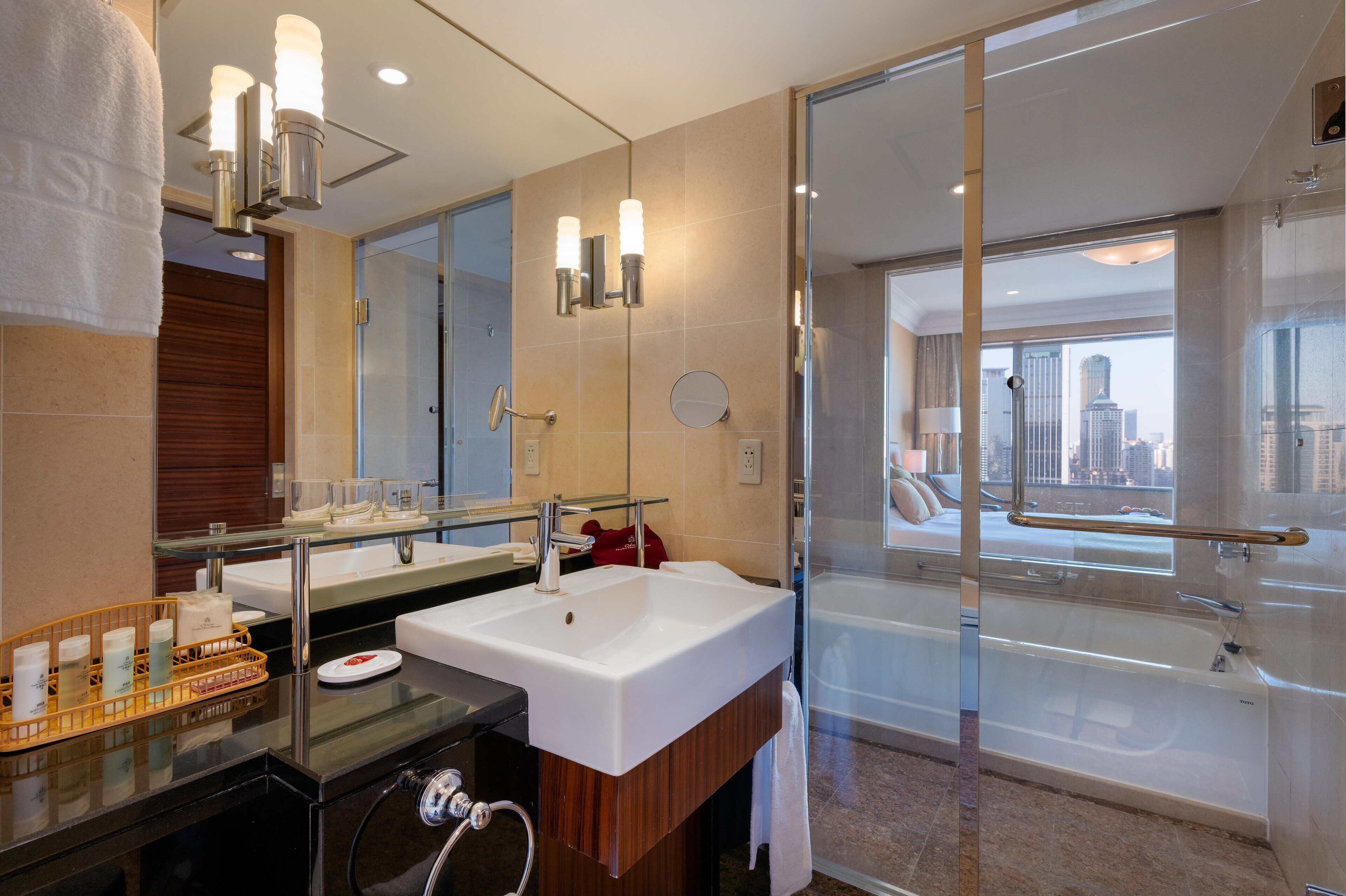 A modern bathroom with a large mirror, dual sinks, a glass-enclosed shower, and a city view.