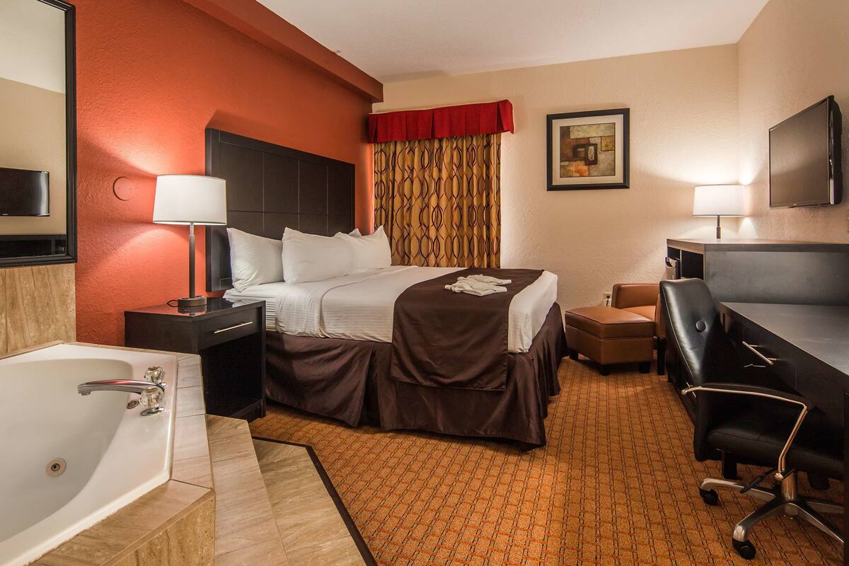 A hotel room with a bed, a desk, a chair, a desk lamp, a TV, a bathroom with a bathtub, and a shower.