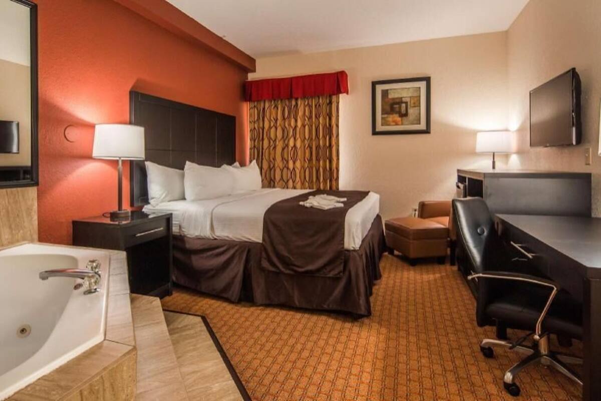 A hotel room with a bed, desk, chair, TV, lamp, and a large bathtub.
