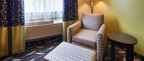 Una habitación de hotel con un sillón estampado, una mesita pequeña, un tapete decorado y una ventana con cortinas.